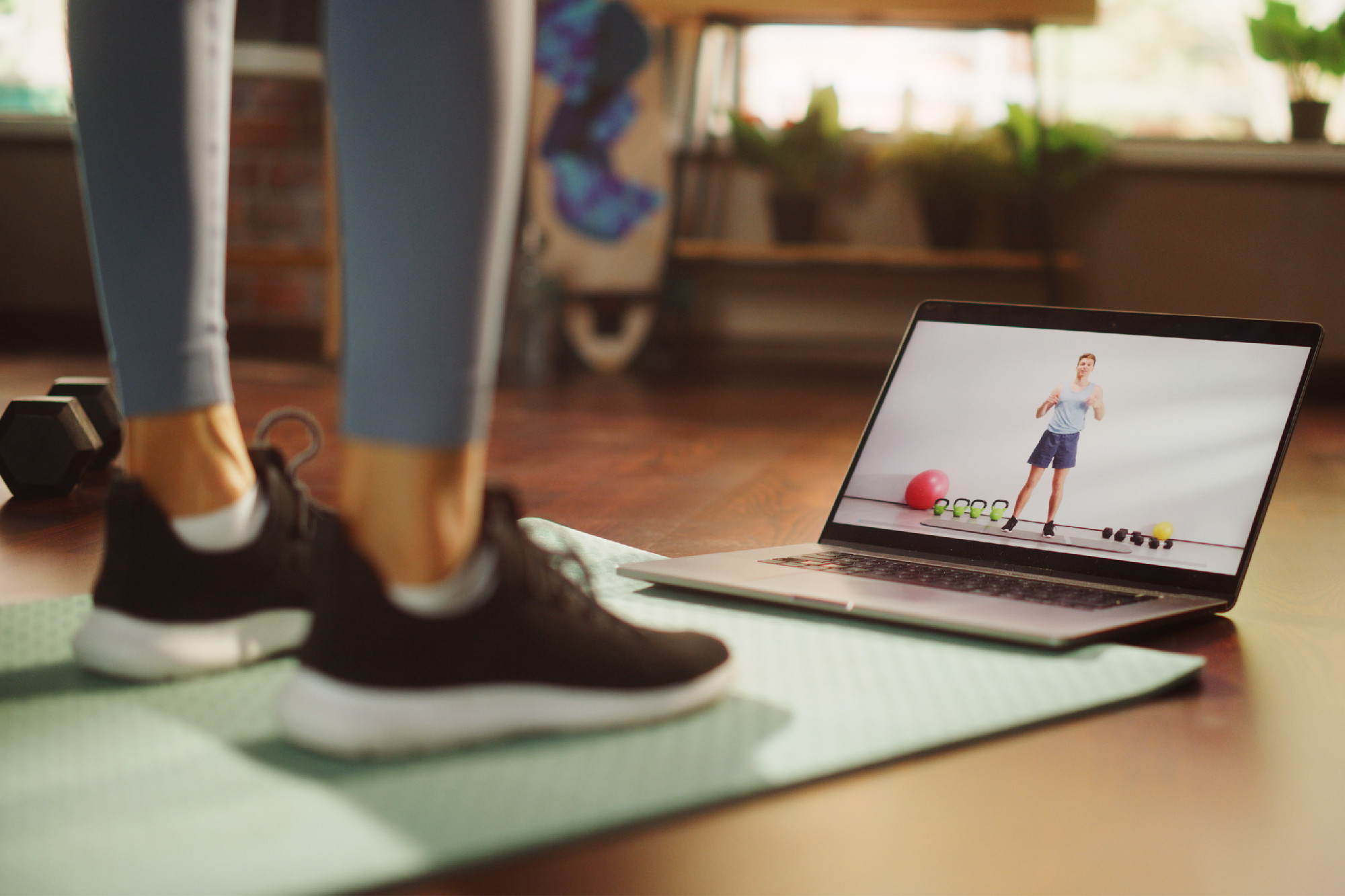 Woman standing on mat with laptop on floor, showing exercise routines